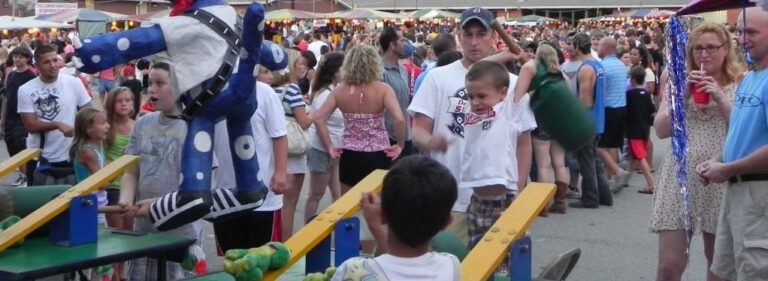 Children play a carnival game where they hit a see-saw with mallets to launch a dummy into the air. A crowd gathers, enjoying this fun idea perfect for summer church activities. In the background, colorful tents and booths showcase various items.