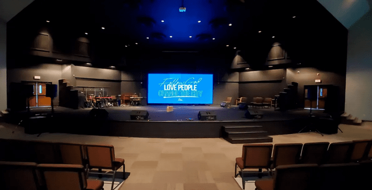 The interior of the church auditorium showcases a stage with musical instruments and rows of empty brown chairs. A large screen on the stage displays "Experience God, Love People, Celebrate Life" in blue and white, highlighting the church's transformative online experience.