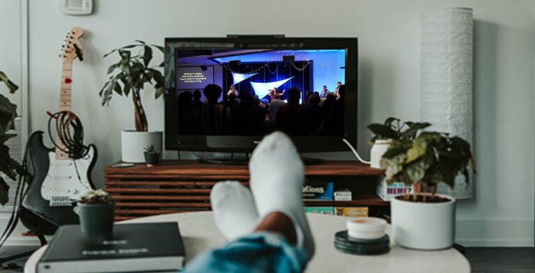 A person with white socks rests feet on a coffee table, streaming their musical journey of a concert on TV. The room, filled with plants, books, and a guitar, creates a cozy and guided atmosphere.