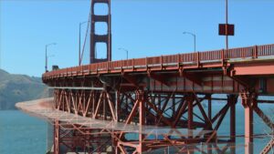 A close-up view of the Golden Gate Bridge, entwined with AI-driven suicide prevention technology, showcases its iconic red-orange structure against clear blue skies. The bridge majestically spans over the water, with distant hills visible on the left.