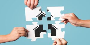 Four hands holding puzzle pieces that fit together to form the silhouette of a house on a light blue background symbolize the careful alignment in church mergers, considering do’s and don'ts for successful collaboration.