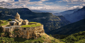 An ancient stone church sits atop a rocky cliff, surrounded by lush green mountains and deep valleys under a partly cloudy sky.