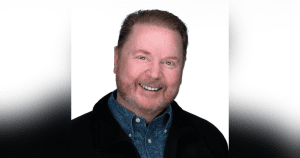 A smiling middle-aged man with reddish hair and beard, wearing a blue patterned shirt and a dark jacket, poses against a plain white background—an author known for his inspiring Christian fantasy novels.