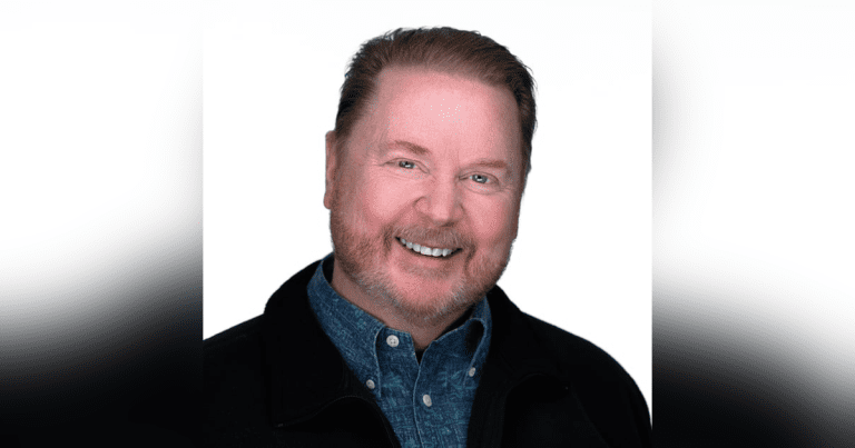 A smiling middle-aged man with reddish hair and beard, wearing a blue patterned shirt and a dark jacket, poses against a plain white background—an author known for his inspiring Christian fantasy novels.