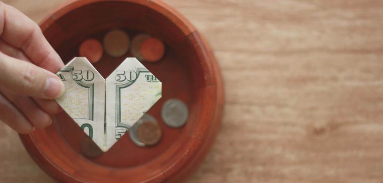 A hand holds a fifty-dollar bill folded into a heart shape above a wooden bowl of assorted coins, illustrating Church stewardship on a light brown surface.
