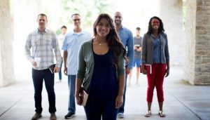 A group of five young adults stand in a row outdoors, smiling at the camera; the woman in front holds a book, while the others stand slightly behind her with relaxed, cheerful expressions.
