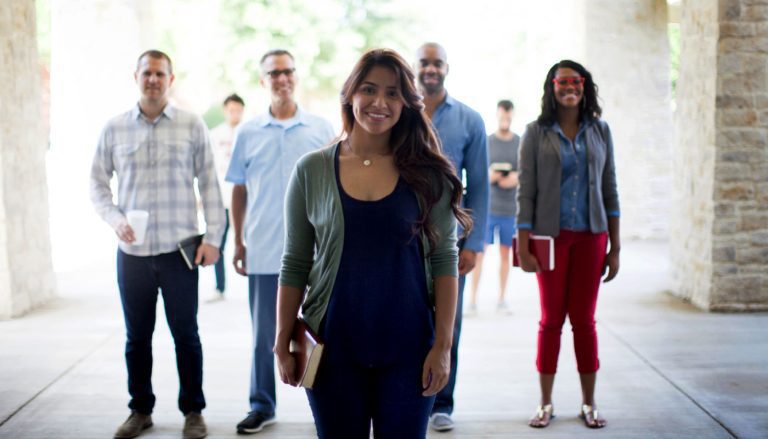 A group of five young adults stand in a row outdoors, smiling at the camera; the woman in front holds a book, while the others stand slightly behind her with relaxed, cheerful expressions.