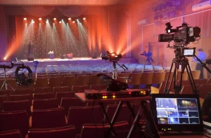 Concert hall with rows of empty seats, stage lights shining on a curtained stage, and several video cameras set up for filming or broadcasting an online church event to boost future engagement.