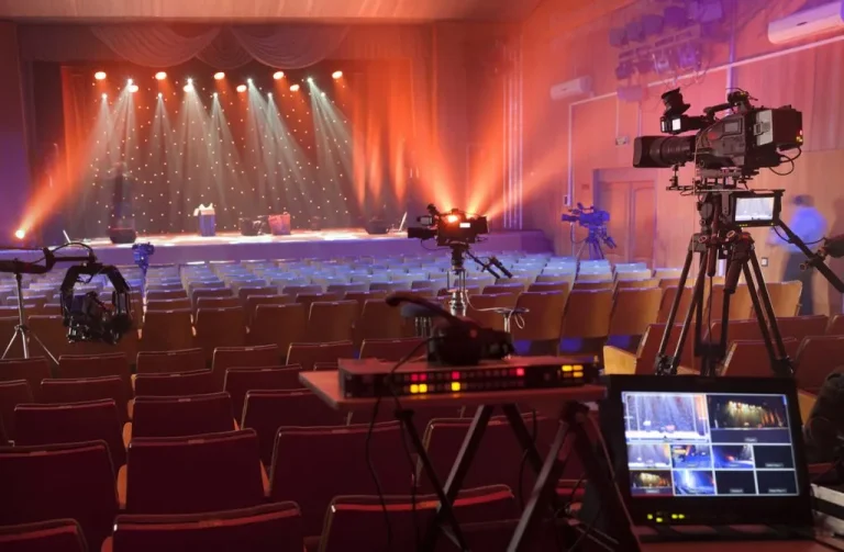 Concert hall with rows of empty seats, stage lights shining on a curtained stage, and several video cameras set up for filming or broadcasting an online church event to boost future engagement.