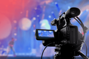 A close-up of a video camera recording a live event on stage with colorful, blurred lights and a person performing in the background—perfect for streaming video for churches.