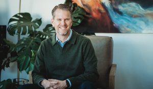 Kenny Wyatt, a smiling man with short light brown hair and a beard, sits in an armchair wearing a dark green sweater over a collared shirt. Large green plants and a colorful abstract painting highlight his people-first ministry setting.