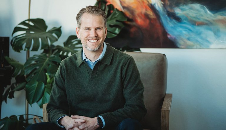 Kenny Wyatt, a smiling man with short light brown hair and a beard, sits in an armchair wearing a dark green sweater over a collared shirt. Large green plants and a colorful abstract painting highlight his people-first ministry setting.