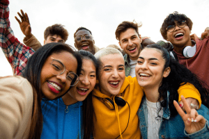 A diverse group of Gen Z and Millennials smiling and posing closely together for a selfie outdoors, some with headphones and casual clothes. One person flashes a peace sign. The mood is cheerful and friendly.