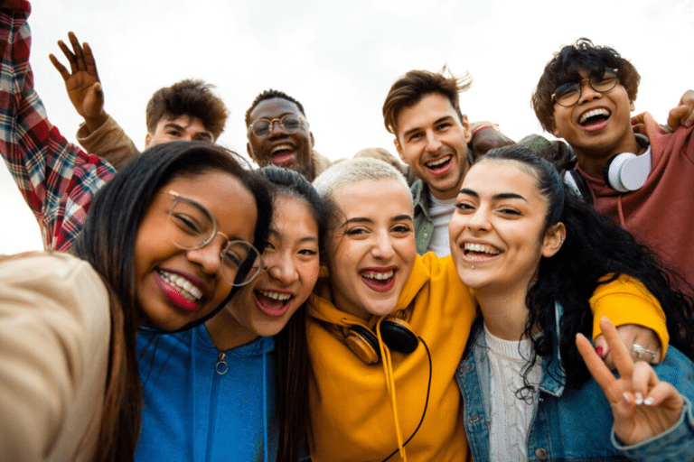 A diverse group of Gen Z and Millennials smiling and posing closely together for a selfie outdoors, some with headphones and casual clothes. One person flashes a peace sign. The mood is cheerful and friendly.