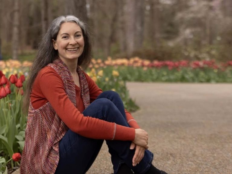 A smiling woman with long gray hair sits on a path in a park, surrounded by blooming red and yellow tulips. Radiating positivity, she embodies a hopeful path amid nature's beauty, wearing a red top, patterned vest, and blue jeans.