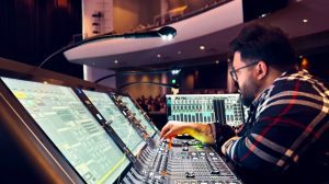 A person wearing glasses and a plaid shirt operates a large audio mixing console with multiple screens and controls in a modern auditorium, managing the complexities of FOH vs Broadcast sound for an AI Mix Church service.